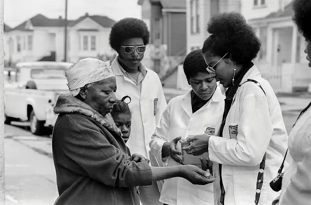 A group of Black women in lab coats helping an older Black woman and a child in the middle of the street.