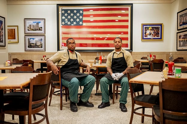 Two men in work clothes sit in front of the American flag at a restaurant.