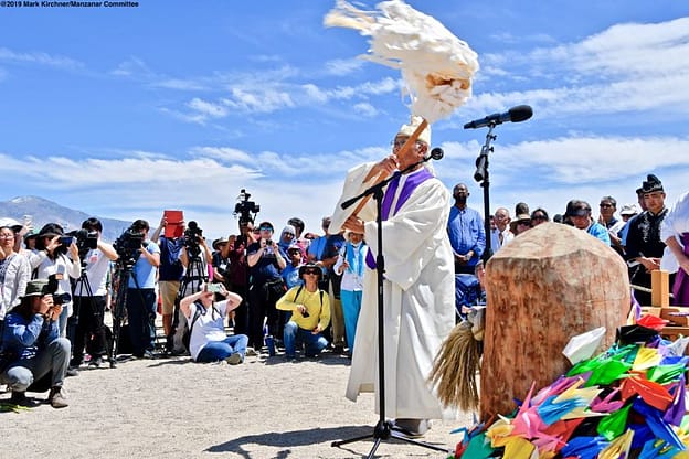 A festival, filled with people. One prominent figure wears headdress and white garb.