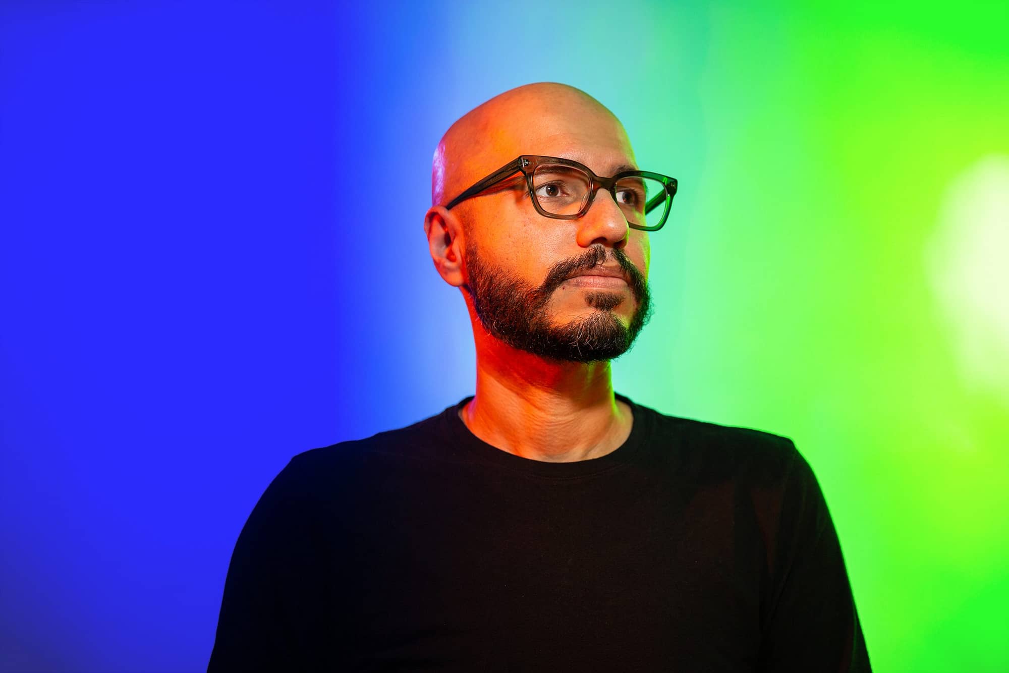 Safwat is dressed in a black top, wearing glasses with dark frames. He stands across a background that is vibrantly lit with blue and green. He is looking up towards his left, so that we are looking at him while he is looking into the distance.