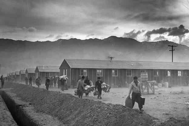 A black and white photo of Japanese internment; rows of housing and people with luggage